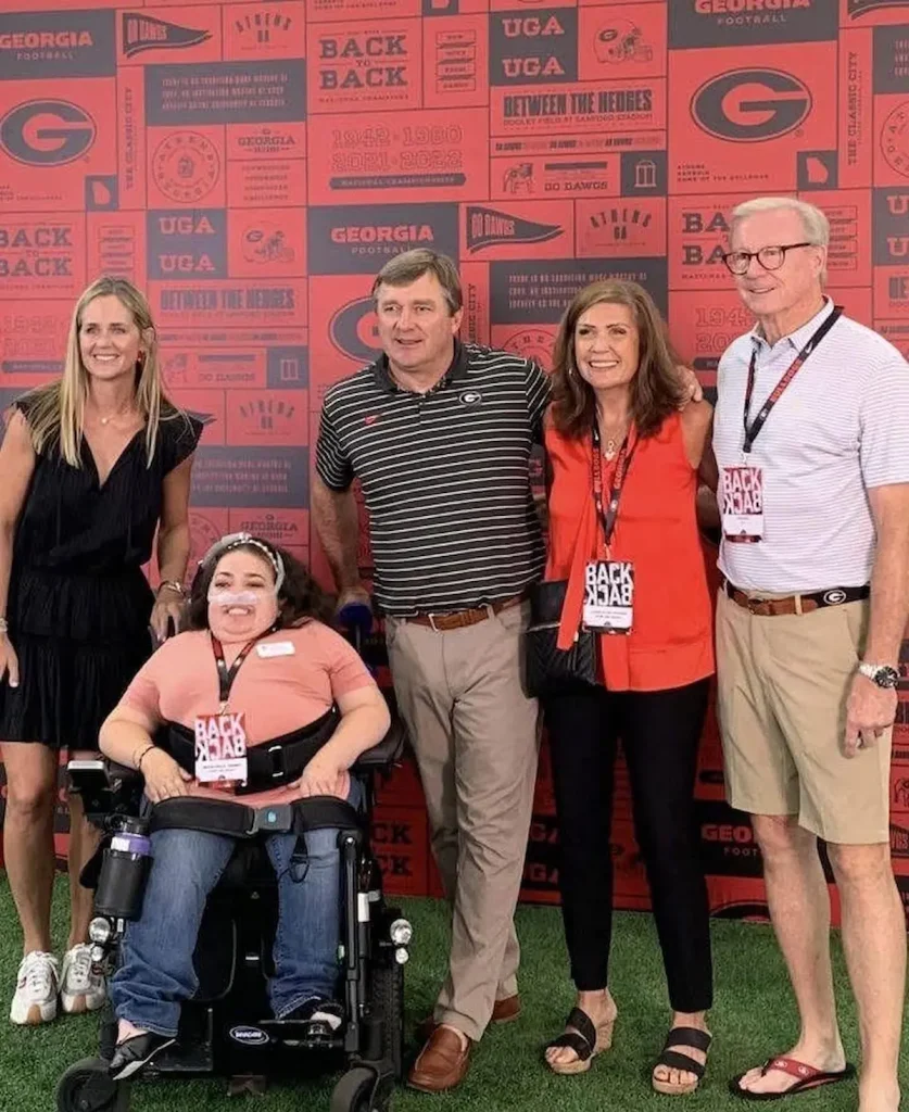 Five people, four standing and one seated in a wheelchair, are posing for a photo in front of a red wall adorned with various sports-related logos and text, including "GEORGIA FOOTBALL" and "BACK BACK BACK". From left to right, a blonde woman in a black dress stands smiling. Next to her, a person with a face mask, possibly in a wheelchair, smiles at the camera. In the center, a man in a striped polo shirt and khaki pants stands with his arm around the smiling woman to his right, who is wearing an orange top and black pants. On the far right, a man in a striped polo shirt and khaki shorts smiles, wearing glasses and sandals. All individuals appear happy and are looking towards the camera.