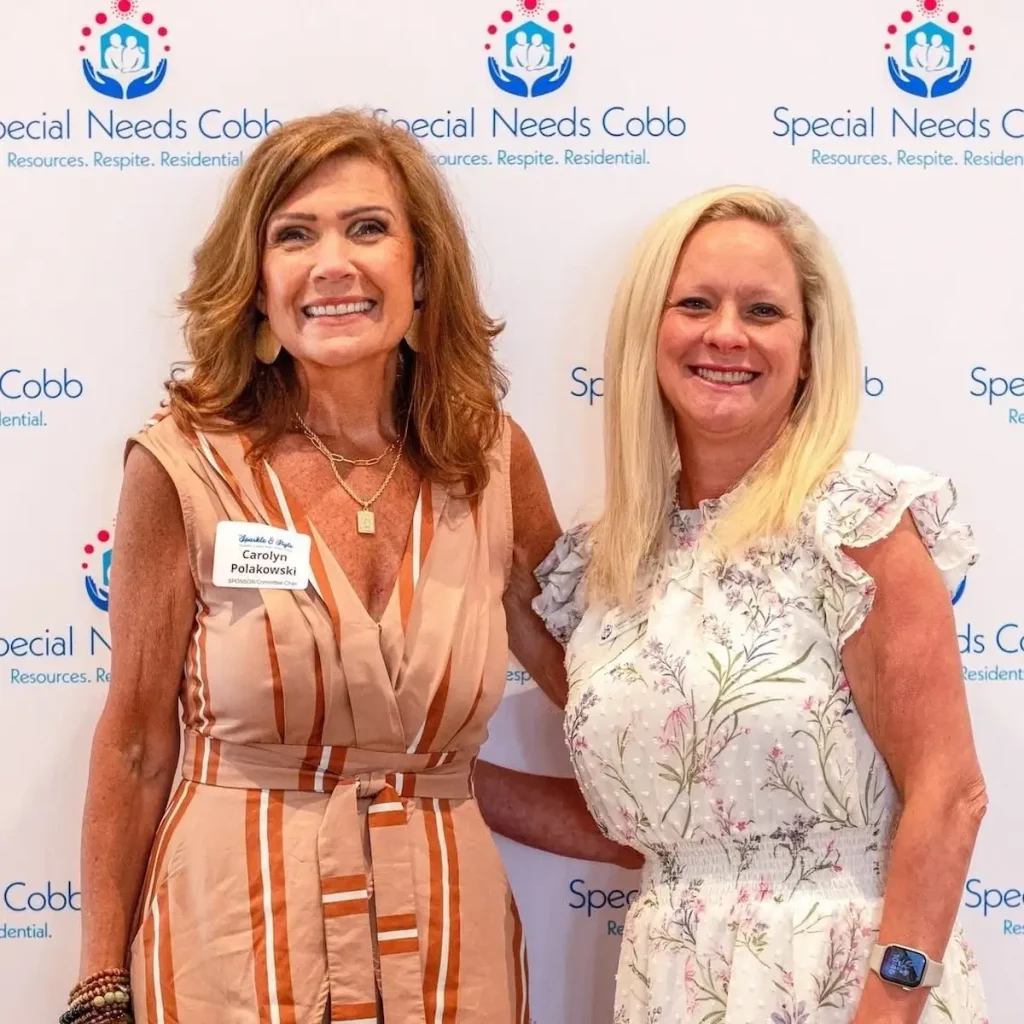 Two smiling women are standing side-by-side in front of a white wall with blue and pink logos and text. The woman on the left has long, wavy brown hair, wears a sleeveless striped dress with a tie-waist, and has a nametag that reads "Carolyn Polanowski." She is also wearing necklaces and bracelets. The woman on the right has long blonde hair, wears a white floral sleeveless dress with ruffles, and has a watch on her left wrist. Both are looking at the camera. The text on the wall includes phrases like "Special Needs Cobb" and "Resources. Respite. Residential."